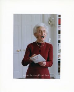 Color candid photograph of Hortense Hirsch. She wears a red dress and a pearl necklace. She's opening an envelope and laughing.