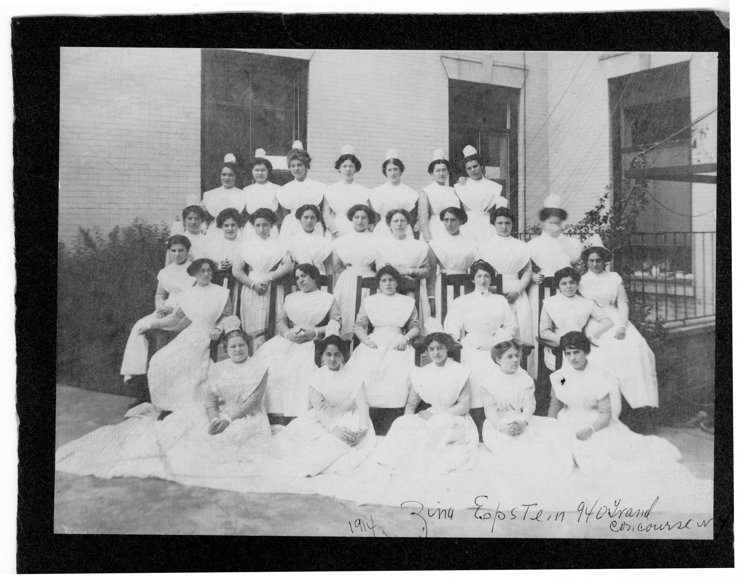 A black and white photograph from 1914. Four rows of young women in nursing uniforms, composed of floor length white dresses and a starched cap, pose for a class photograph in front of the Beth Israel Hospital Jefferson and Cherry Streets location. Handwriting at the bottom reads "1914 - Zina Epstein 940 Grand Concourse"