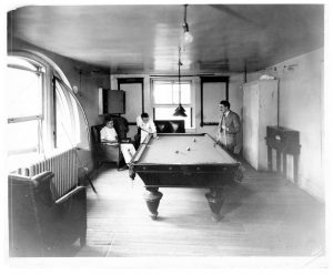 A black and white photograph circa the 1920s. Two members of the Beth Israel Hospital house staff play pool in the house staff lounge at the Jefferson and Cherry Street location. A third doctor, sitting in an armchair, watches them play.