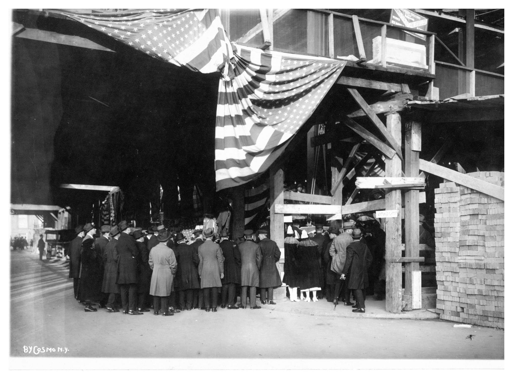 Black-and-white photograph of a crowd with their back turned to the camera. They stand under an American flag at the Dazian Pavillion construction site watching the cornerstone being laid.