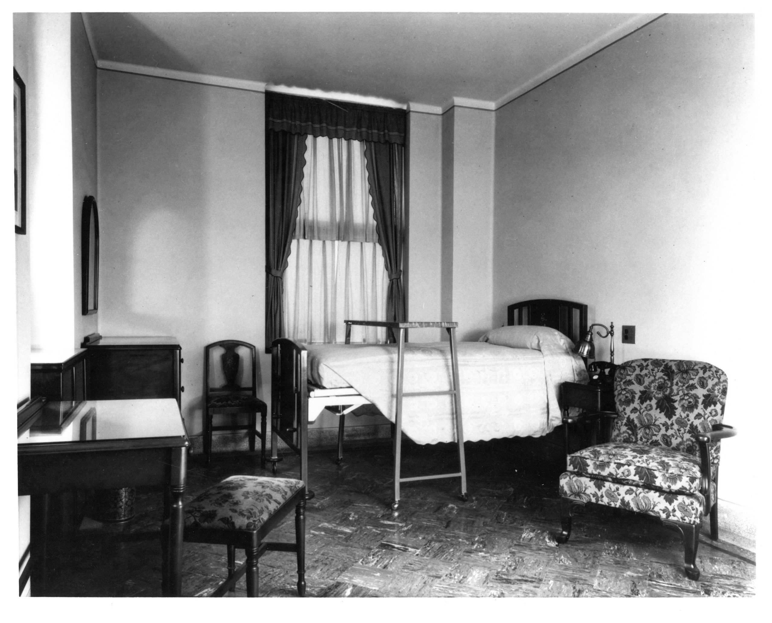 Black-and-white photograph of patient room. Furniture includes twin bed with tray, armchair, dresser, desk with chair. There's also a window with a curtain.
