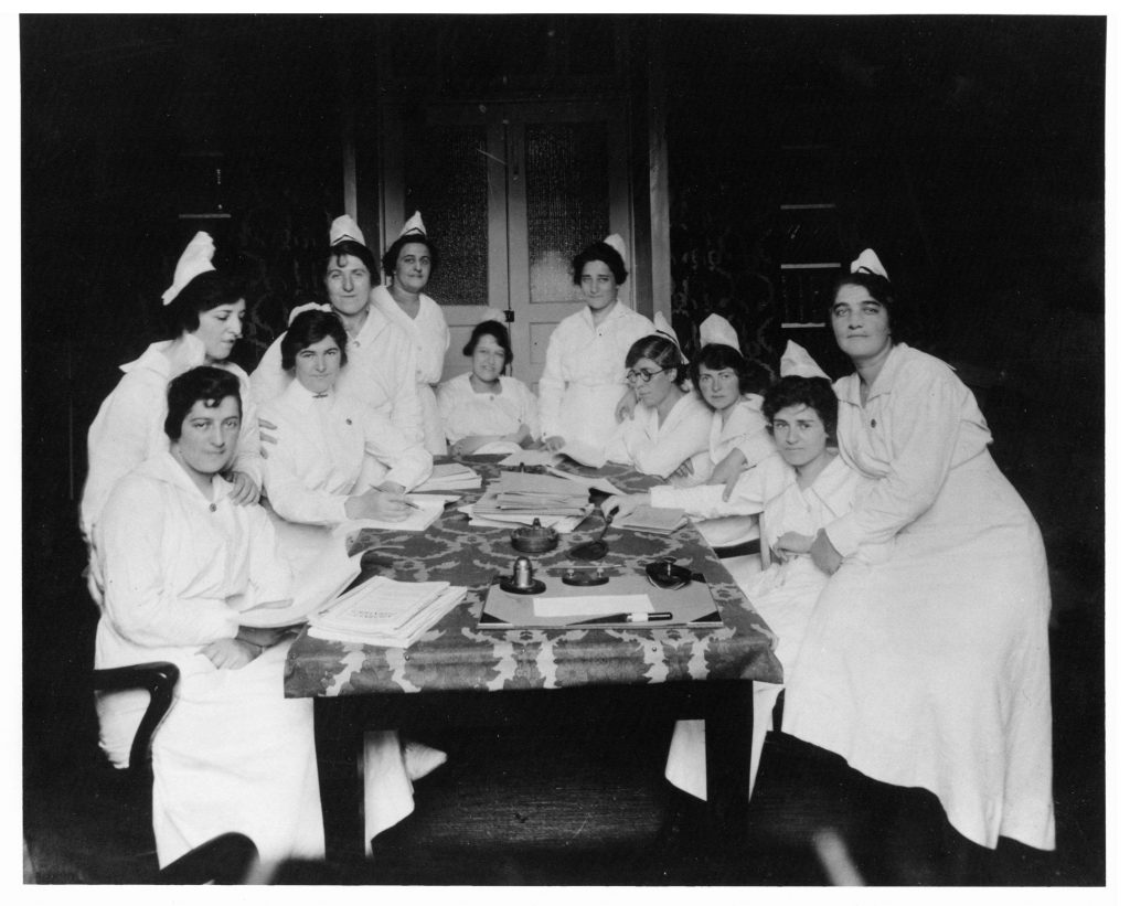 Black and white photograph of eleven nursing students in uniform studying around a large table in a dark room