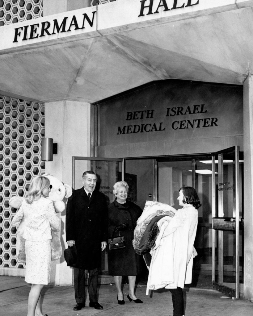 Harold and Minnie Fierman stand between two Phillips School of Nursing Students carrying clothes and a teddy bear in front of Fierman Hall